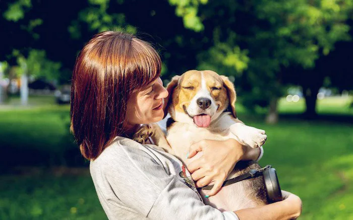 Fotografía de mujer cargando un perro sonriente y feliz en un parque.
