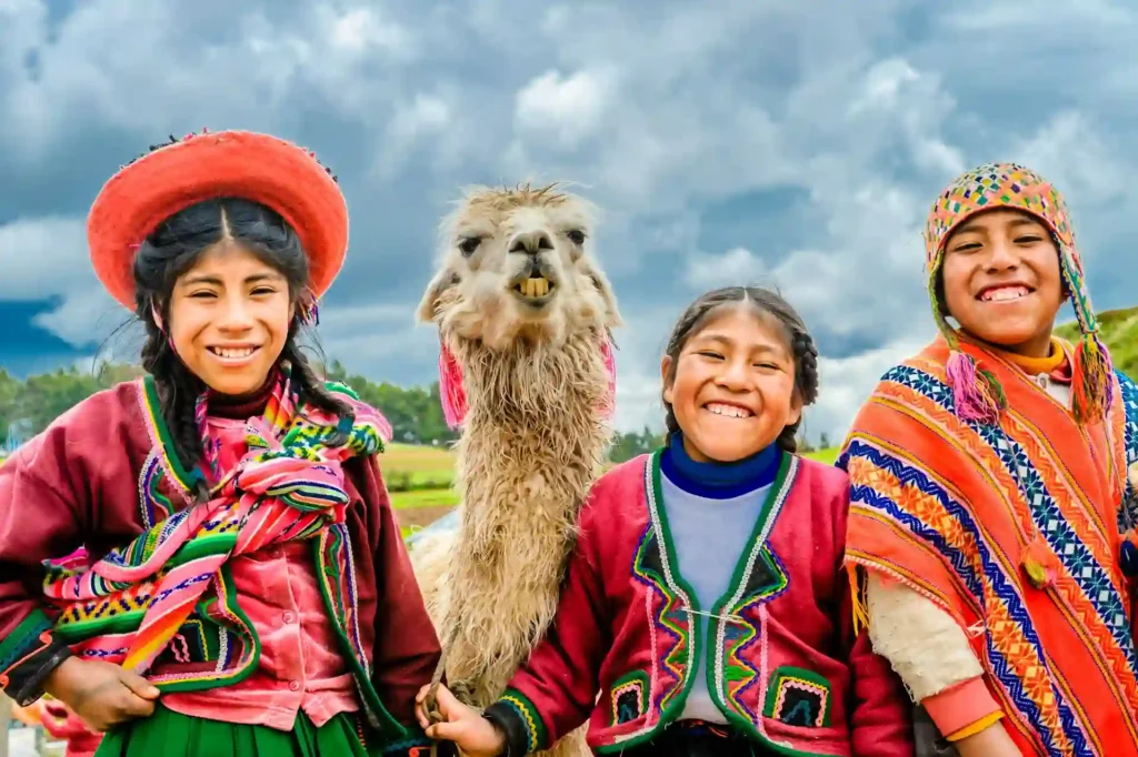 Grupo de niños con vestimenta tradicional peruana posando con una llama en un paisaje montañoso, representando la conexión cultural y las experiencias auténticas del turismo transformador en Perú.