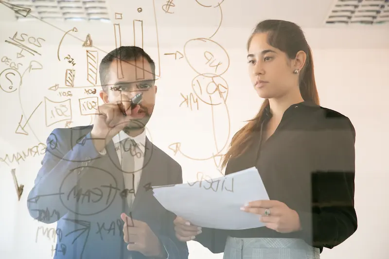administrador-escribir-grafico-sobre-placa-de-vidrio-colega-bastante-femenina-joven-profesional-que-sostiene-el-documento-y-que-mira-el-grafico-en-la-sala-de-conferencias-concepto-de-marke